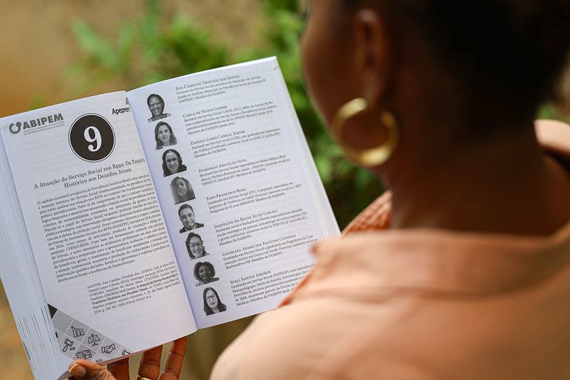 Uma pessoa segura um livro aberto sobre Regimes Próprios de Previdência Social (RPPS), focado na página de autores da ABIPEM. A pessoa usa brincos de argola dourados e uma camisa de linho coral. O fundo é desfocado.