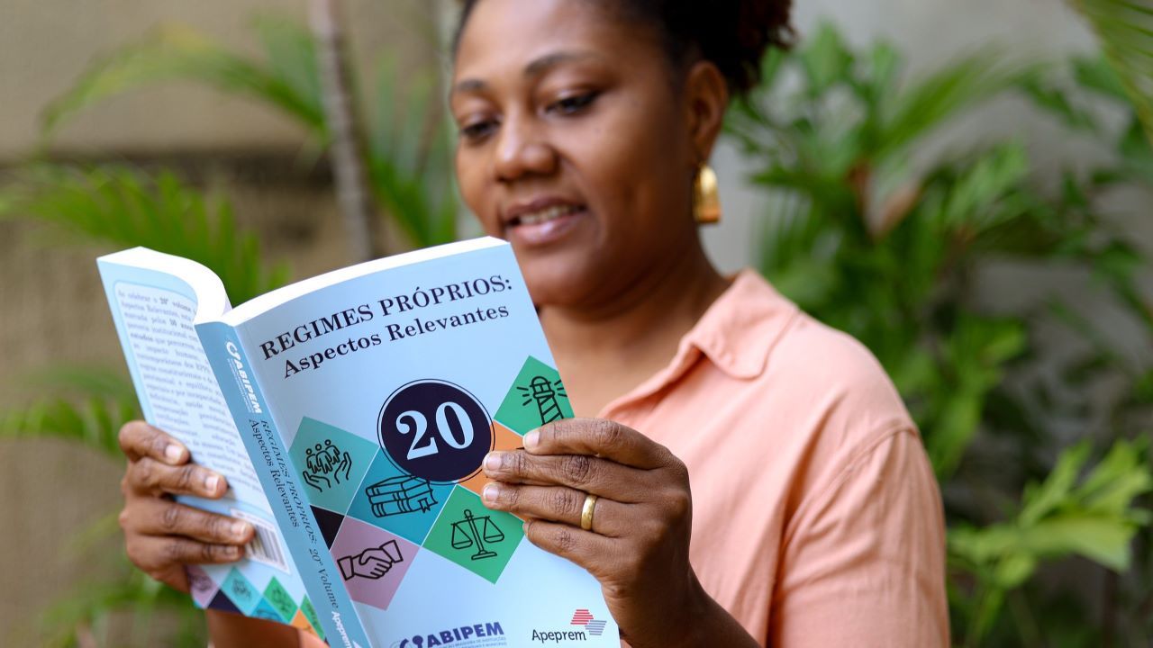 Mulher lendo livro com capa azul em um ambiente externo com plantas ao fundo.